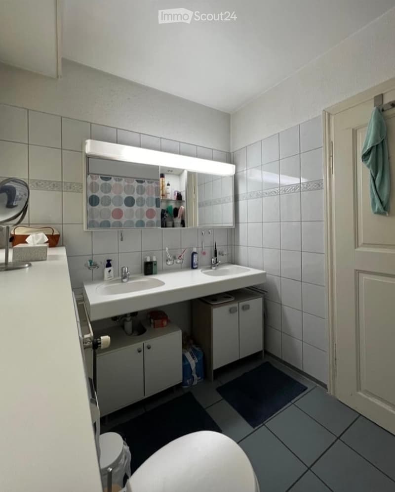 A modern bathroom with a double sink vanity, large mirror with built-in storage, and tiled walls. The vanity includes cabinets and drawers, and a towel is hanging on the door.