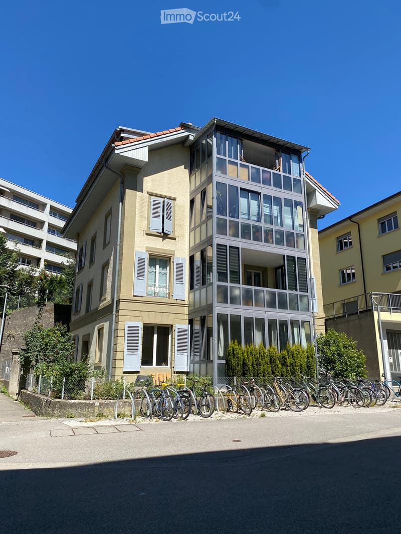 A modern multi-story building with a combination of beige and glass facades, featuring large windows and balconies. Several bicycles are parked in front of the building.