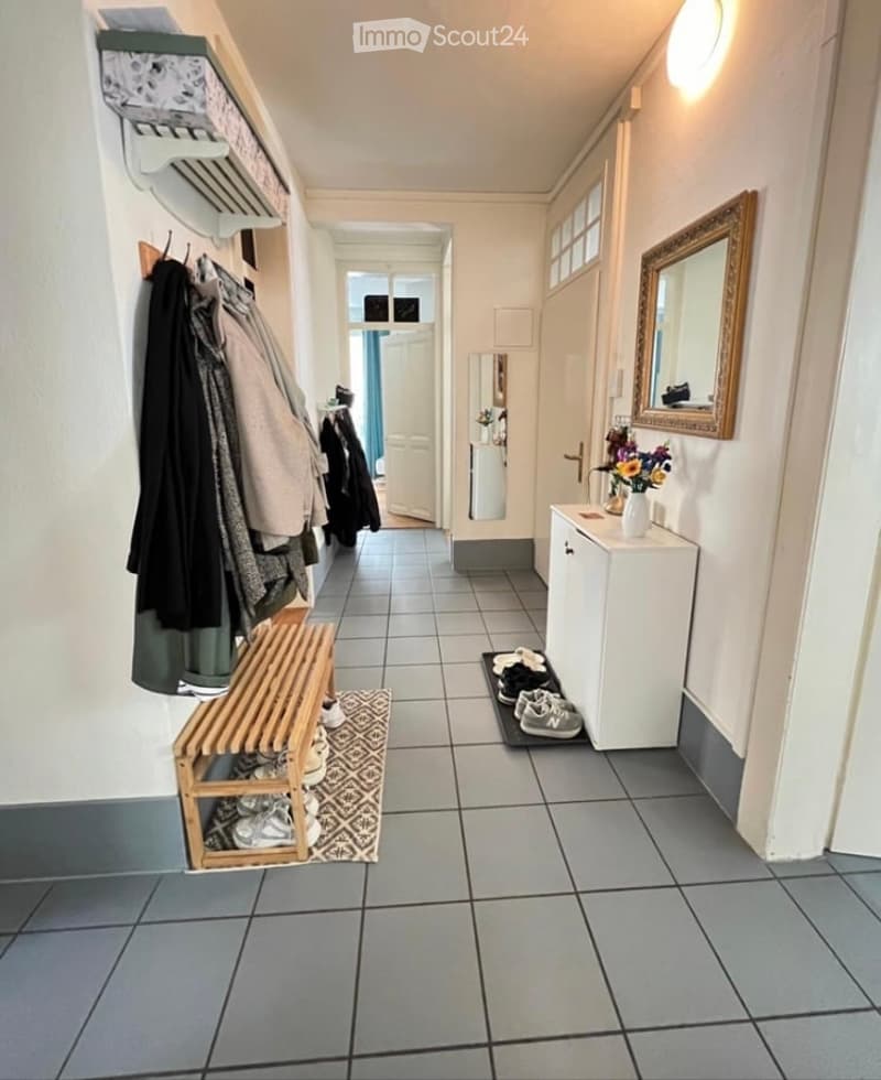 Clean, well-lit hallway with tiled flooring, coat rack, shoe bench, small cabinet, and mirror. The hallway leads to another room with a visible door.