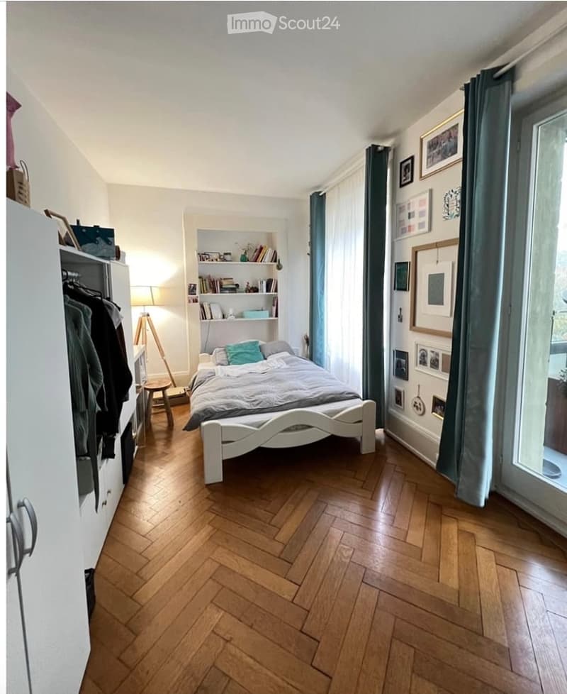 Cozy bedroom with wooden herringbone flooring, white bed with grey bedding, built-in wardrobe, bookshelf, and decorative picture frames on the wall.