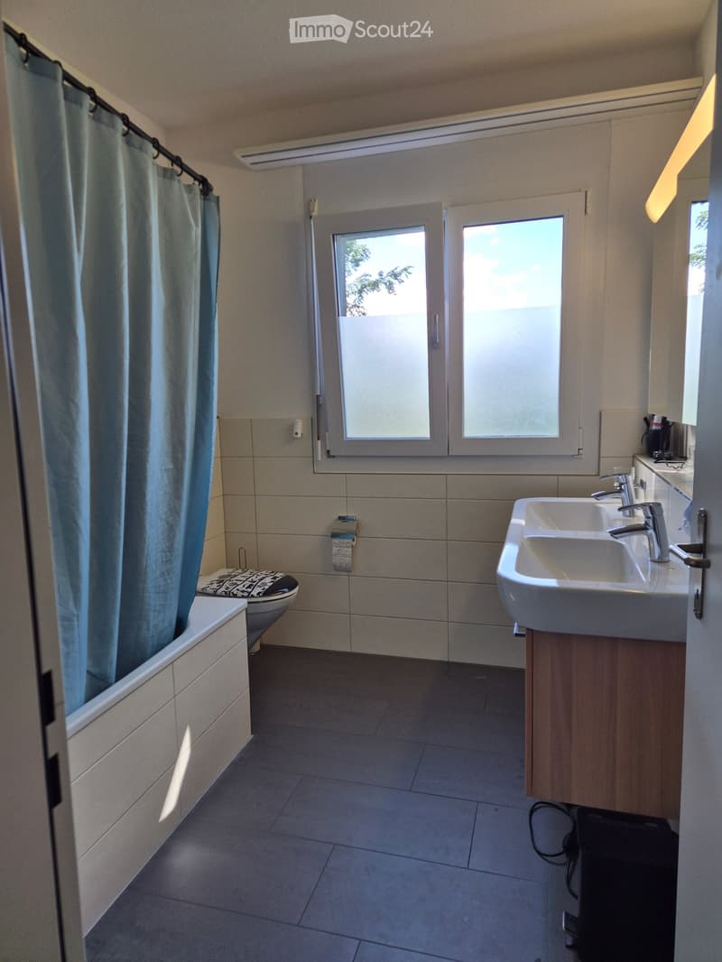 Modern bathroom with a bathtub, toilet, double sink, and large windows. Features tiled walls and floor, a wooden vanity, and a blue shower curtain.