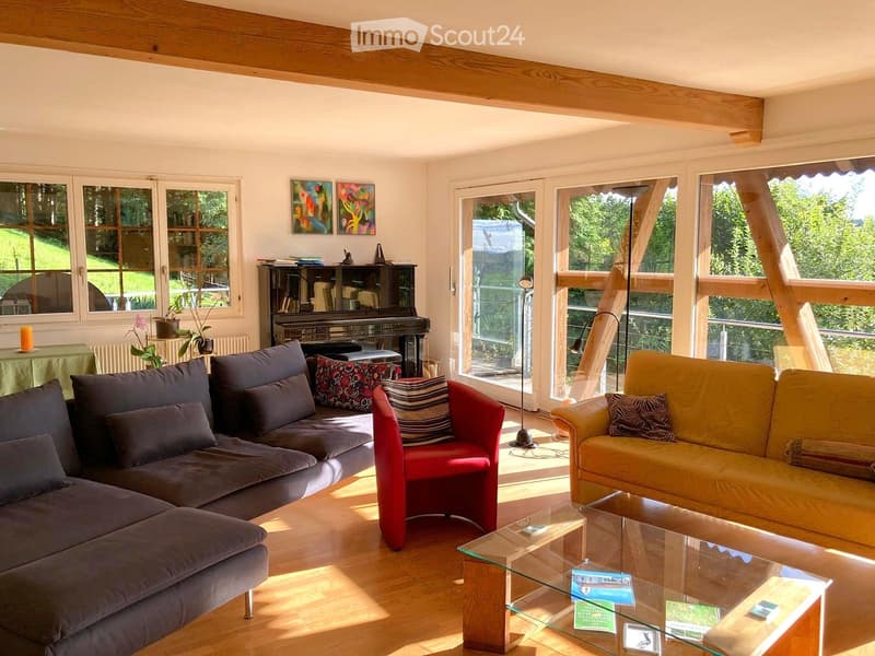 Bright living room with wooden floors, large windows, and a view of greenery. Features a grey sectional sofa, a red armchair, a yellow loveseat, a glass coffee table, a piano, and a small table with a potted plant.