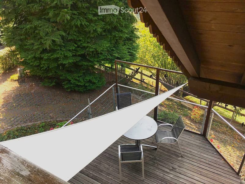 Balcony with wooden decking, a white sunshade, a round table, and two black chairs, overlooking a garden area with trees and a wooden fence.