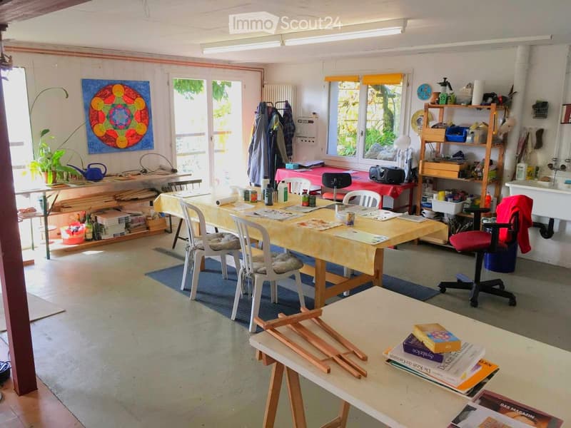 A spacious, well-lit studio with large windows, multiple tables, chairs, shelves filled with art supplies, a painting on the wall, and a sink area.