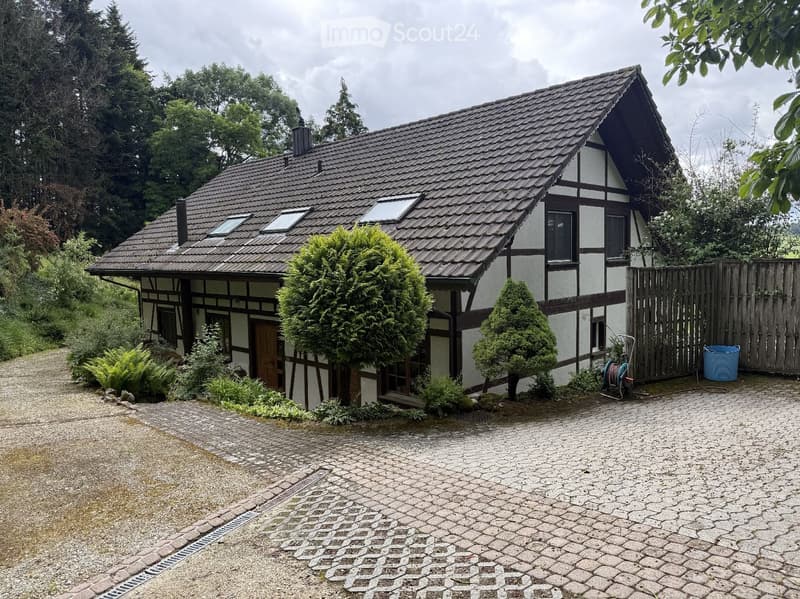 Two-story house with a tiled roof, white and dark wood exterior, surrounded by greenery and a gravel driveway.