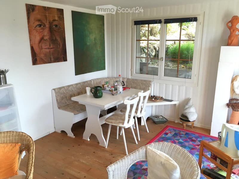 Brightly lit room with wooden flooring, white walls, and large windows. Features a white dining table with a bench seating, several chairs, a wicker chair with an orange cushion, and various decorative items including paintings and sculptures.