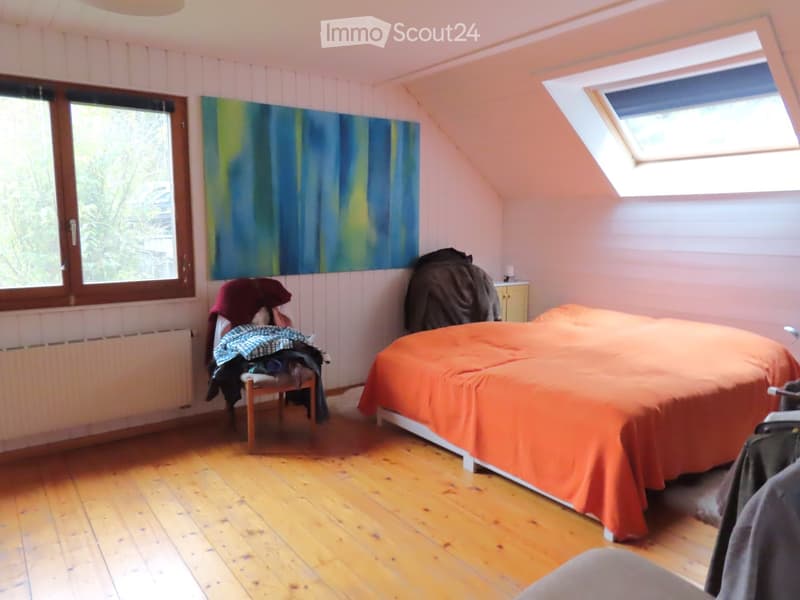 Bedroom with wooden floor, slanted ceiling, large window, skylight, bed with orange cover, chair with clothes, painting on wall, small cabinet.