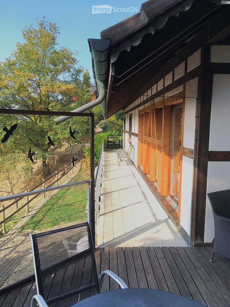 Wooden balcony with metal railing, overlooking a grassy area with trees, featuring a chair and table.