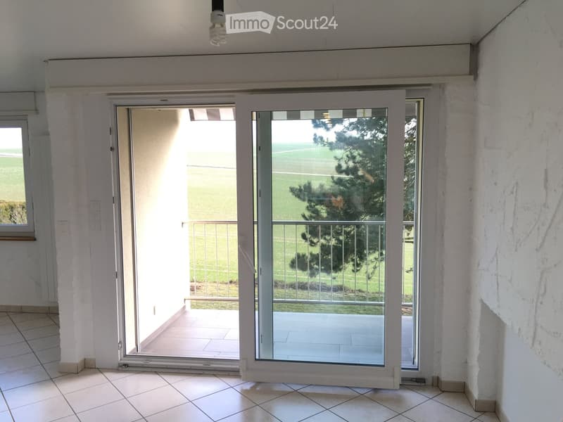 Empty room with tiled floor, white walls, and large sliding glass doors leading to a balcony with a view of a green area and trees.