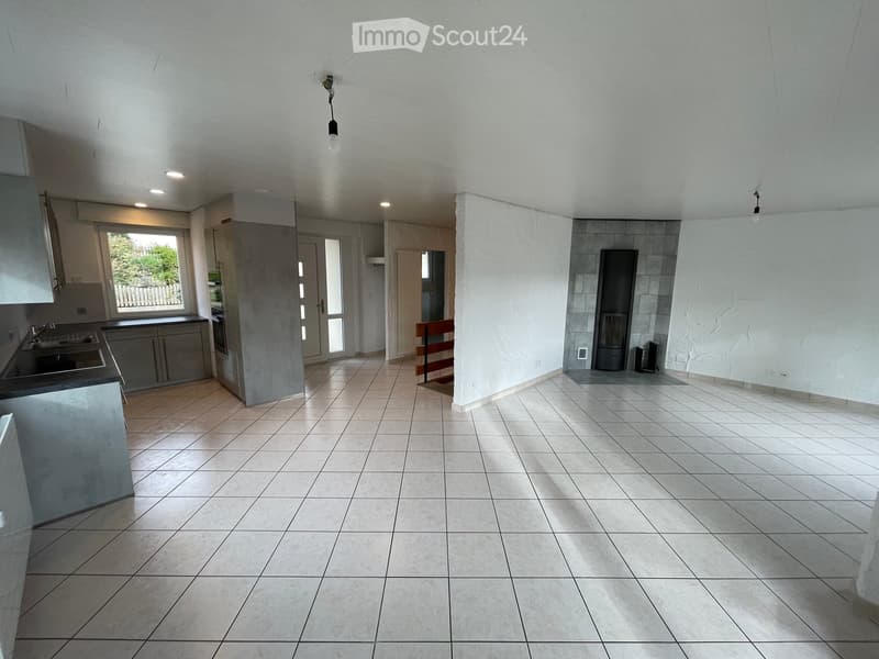 Empty room with tiled floor, white walls, and a small kitchen area with sink and cabinets. There is a doorway leading to another room and a window with a view of greenery outside.