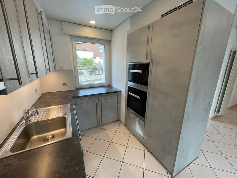 Modern kitchen with stainless steel sink, built-in oven, and grey cabinets. Features tiled flooring and a large window with a view of the garden.