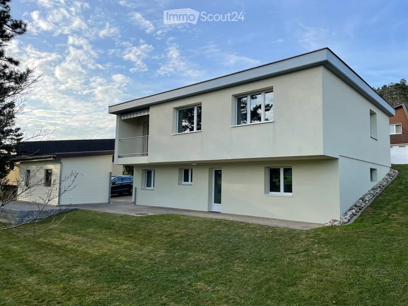 Modern two-story house with light-colored exterior, large windows, and a balcony on the upper level. The house features a garage and is surrounded by a well-maintained lawn.