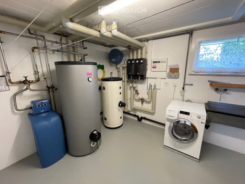 Utility room with a washing machine, water heater, water softener, and control panel, featuring white walls and a window with floral curtains.
