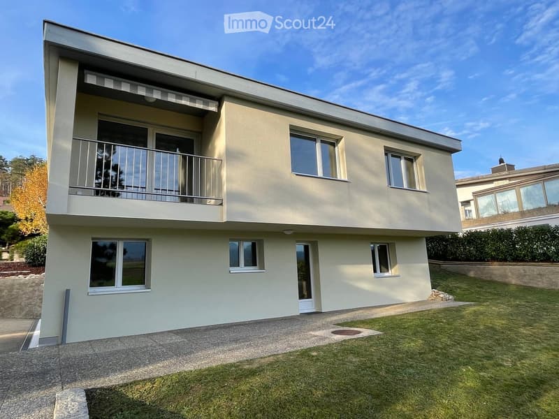 Two-story house with light beige exterior, large windows, and a balcony on the upper level. The house features a well-maintained lawn and a concrete driveway.