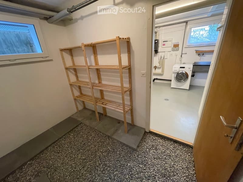 A small utility room with a washing machine, wooden shelves, and a window. The floor is covered with small pebbles.