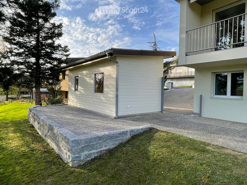 Two-story house with a raised ground floor, white siding, and a balcony on the upper level. The lower level has a concrete patio area with a stone border.
