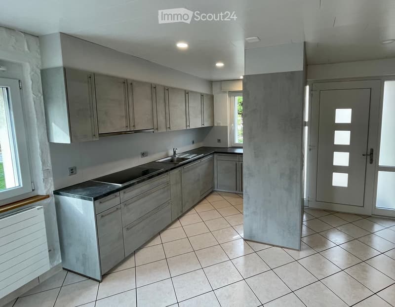 Modern kitchen with grey cabinets, stainless steel sink, and tiled floor. Features include a window with white curtains and a radiator below it.