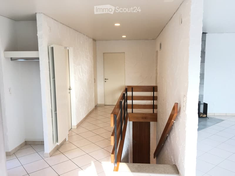 A narrow hallway with tiled flooring, white walls, and a wooden staircase leading upwards. The staircase features a wooden handrail and metal balusters. There is a closed door at the end of the hallway and a partially open door on the left side.