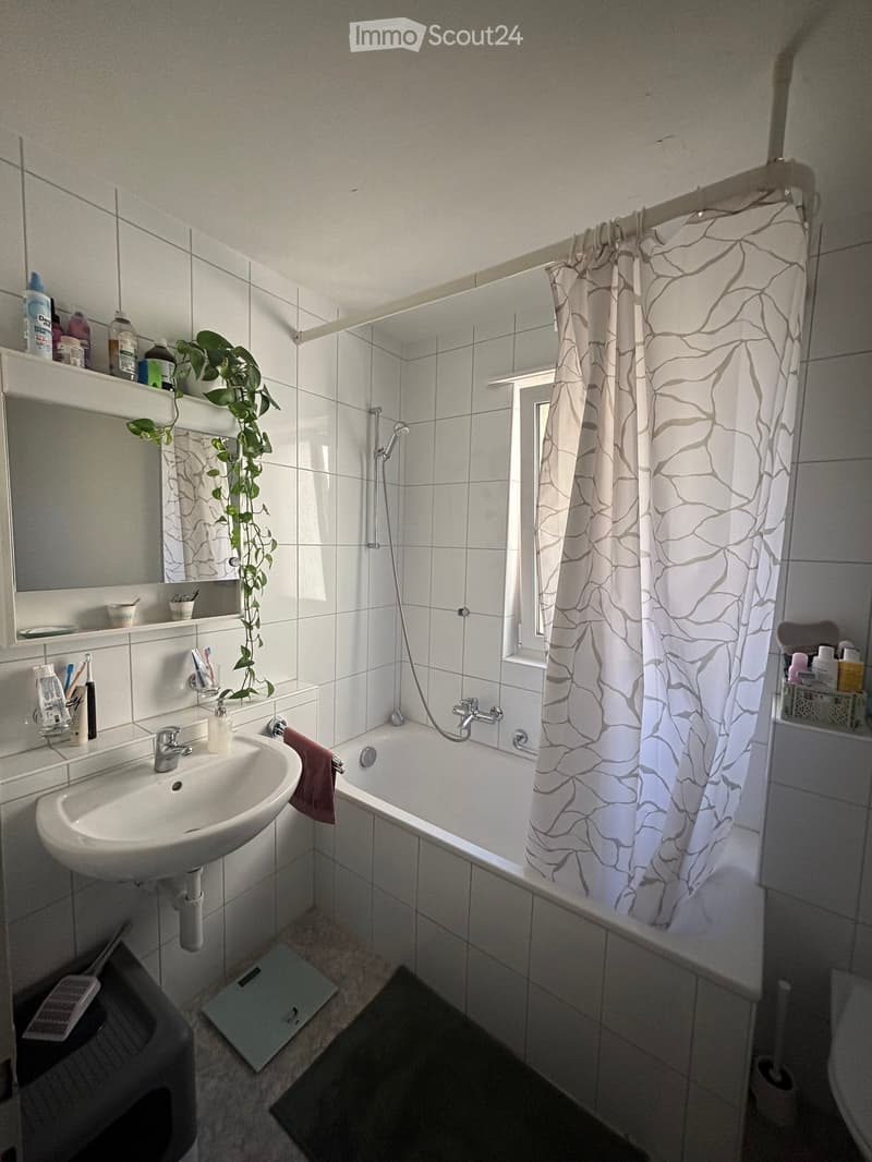 A modern bathroom with white tiled walls and floor, featuring a sink, bathtub with a shower curtain, a mirror, and various toiletries on shelves.