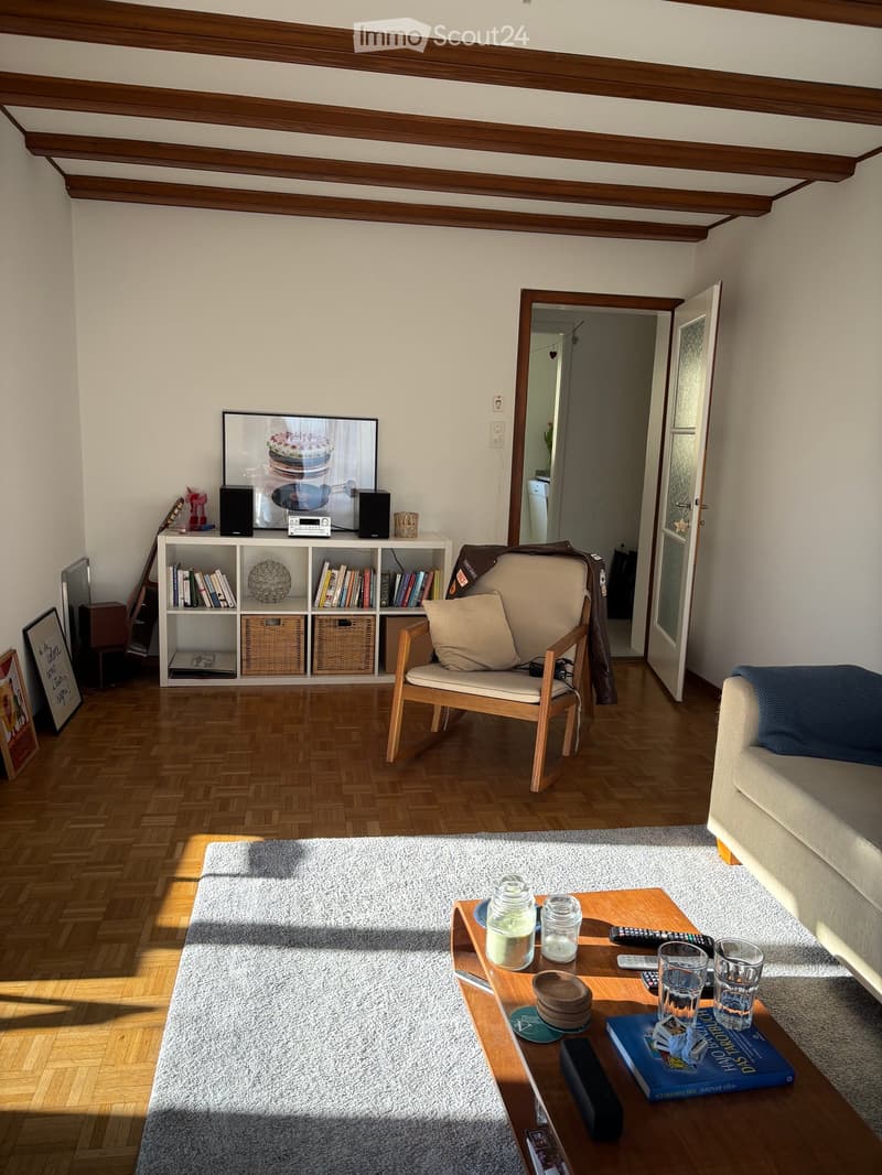 Living room with wooden herringbone flooring, exposed wooden ceiling beams, a cream sofa, a wooden armchair, a coffee table with glass jars and remote controls, a bookshelf with books and decorative items, and a television on a stand.