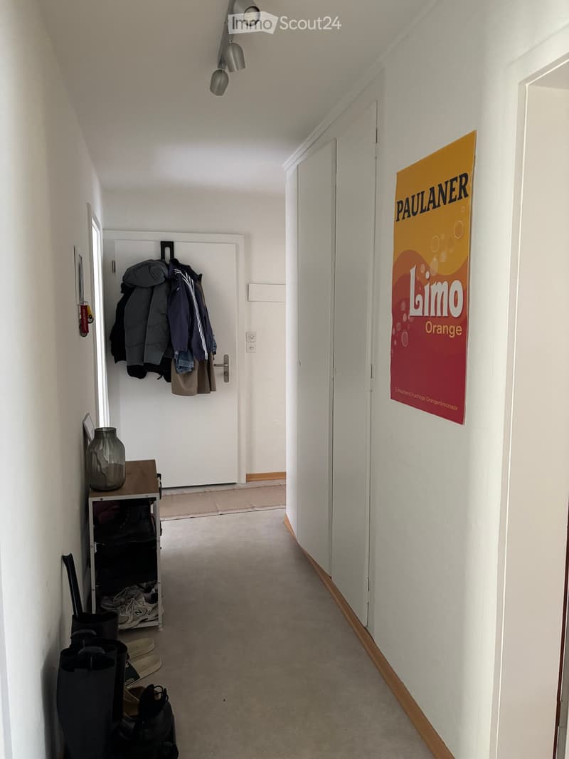 A narrow hallway with white walls, carpeted floor, and a coat rack holding jackets and coats. A poster advertising Paulaner Limo Orange is visible on the right wall.