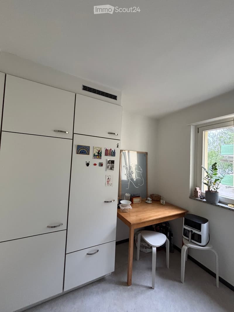 A small, modern room with a refrigerator, a wooden table, a white chair, a small stool, a microwave, a mirror, and a potted plant on the windowsill.