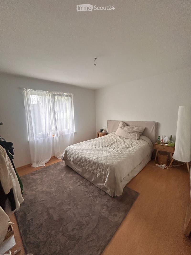 Bedroom with a double bed, nightstand, lamp, and a large window with curtains. The room features a plush rug and wooden flooring.