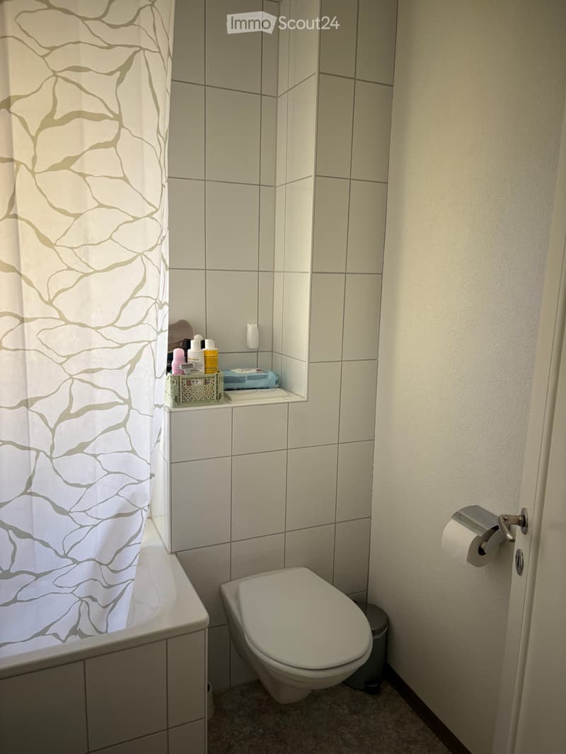 Modern bathroom with a toilet, bathtub, and a shelf holding various toiletries. The walls are tiled, and a patterned shower curtain is visible.