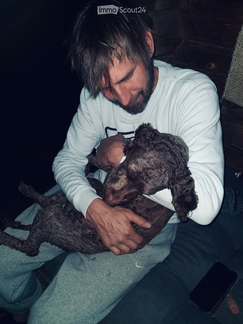 A man in a white shirt is holding and hugging a small, dark-colored dog in a dimly lit indoor setting. The dog appears to be a puppy with a fluffy coat. The man is seated on a couch, and a smartphone is visible on the couch next to him.