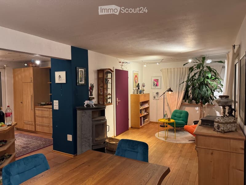 A cozy living room with wooden flooring, a fireplace, a green armchair, a yellow side table, a wooden dining table, and various decorative items including artwork and a plant.