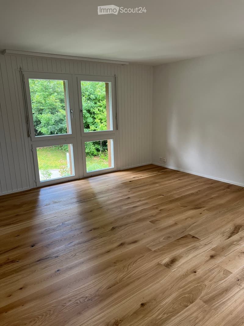 Empty room with wooden flooring, white walls, and large glass doors leading to a green outdoor area.