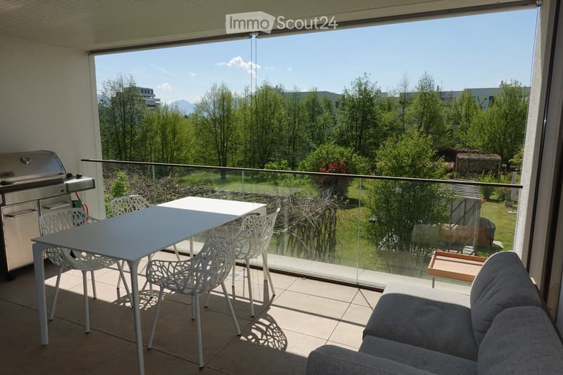 Balcony with a white table, four white chairs, a grey sofa, and a modern grill, overlooking a green landscape with trees and mountains.