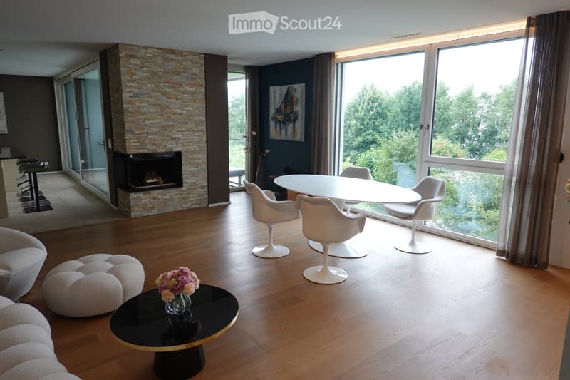 Modern living room with hardwood floors, fireplace, large windows with a scenic view, white dining set, and round coffee table with a flower arrangement.