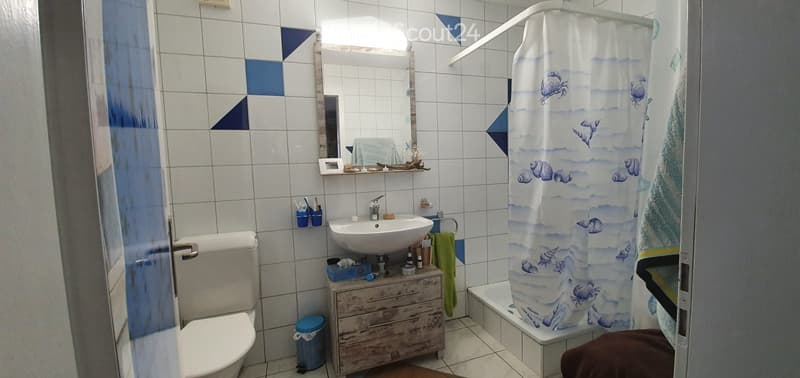 Bathroom with white tiles, a toilet, a sink with a mirror above it, a bathtub with a decorative shower curtain, a wooden cabinet, and a towel hanging on the side.