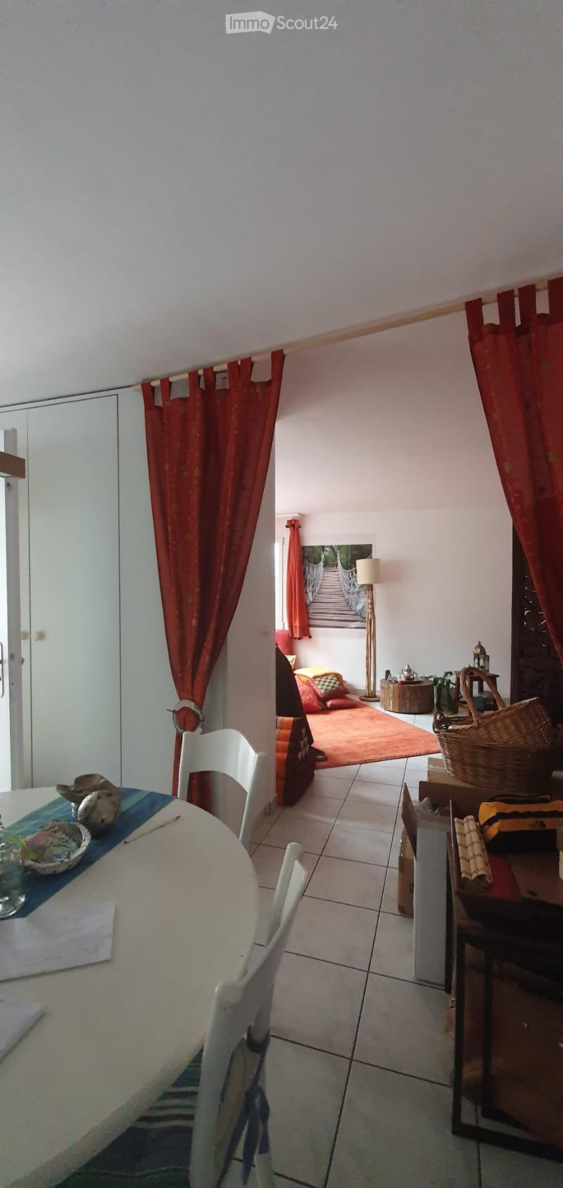 A furnished room with a dining area and a sitting area. The dining area features a round table with chairs, a blue table runner, and decorative items. The sitting area includes a red rug, cushions, a floor lamp, and a small table with a basket and plant.