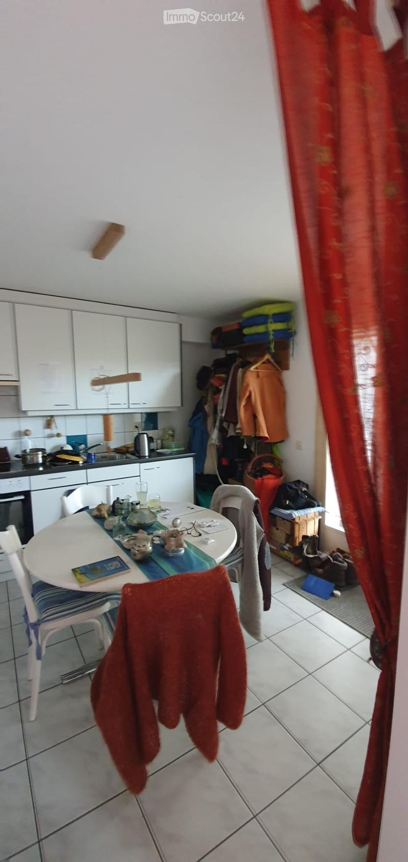 A small kitchen with white cabinets, a dining table with chairs, and various kitchen appliances. There is a red curtain on the window and some clothes hanging on the wall.