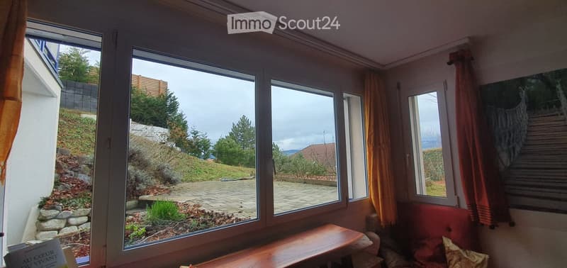 Bright room with large windows offering a scenic view of a landscaped garden and hillside. Features a red sofa, wooden bench, and decorative curtains.