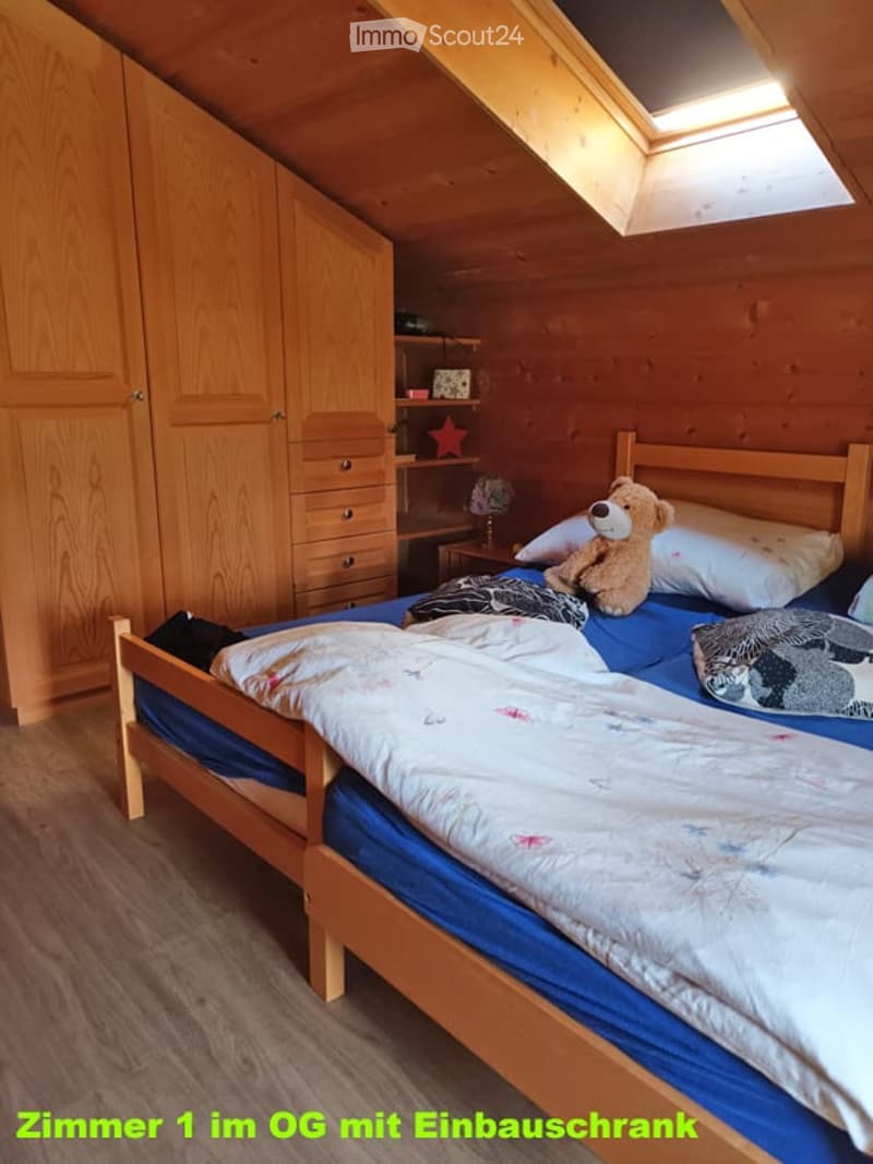 Cozy attic bedroom with wooden walls and ceiling, featuring a single bed with a white comforter, a wooden wardrobe, a small nightstand with a teddy bear, and a shelf with decorative items.
