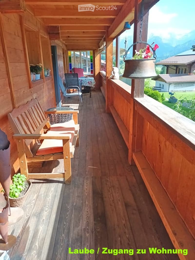 Wooden balcony with a view of mountains, featuring a bench, chairs, a small table, and flower pots.
