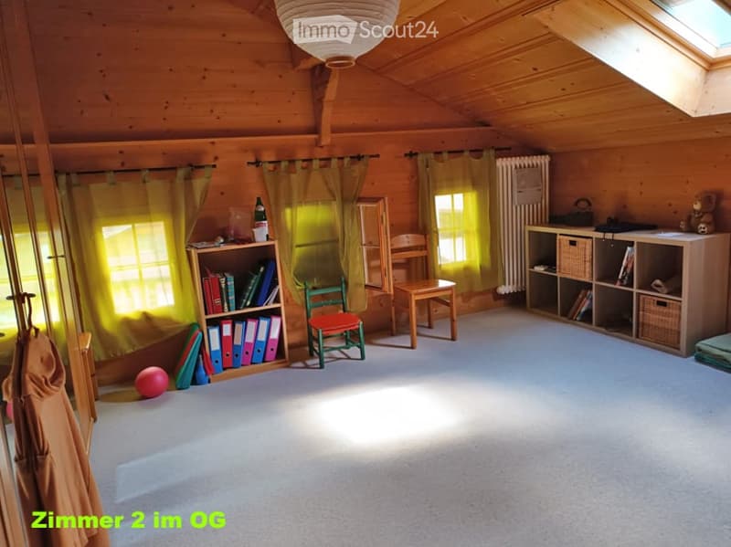 Bright attic room with wooden walls and ceiling, large windows with yellow curtains, a green chair with red seat, a wooden table, a bookshelf with books and binders, a small ball on the floor, and a radiator near the window.