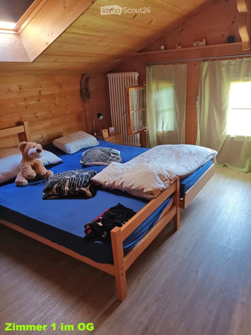 Cozy wooden bedroom with a double bed, multiple pillows, a teddy bear, a nightstand with a lamp, and a radiator. The room features wooden walls and flooring, with a window covered by a curtain.