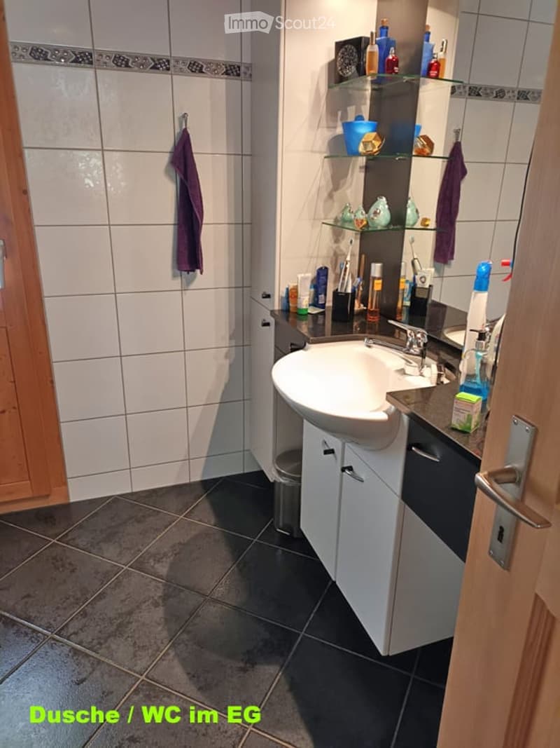 Modern bathroom with white tiles, dark grey floor, white sink, mirror, and various toiletries on the counter. A towel hangs on the wall, and a small cabinet is visible below the sink.