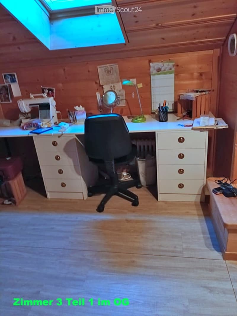 A well-lit room with wooden walls and flooring, featuring a large desk with a chair, a sewing machine, a lamp, a mirror, and a calendar on the wall. The room has a skylight providing natural light.