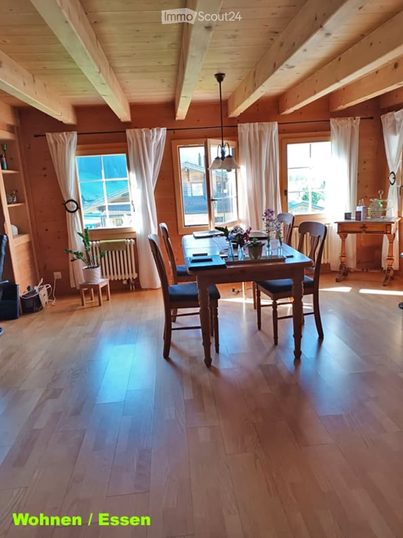 Bright dining room with wooden walls and ceiling, large windows with white curtains, a dining table with chairs, a flower vase, a side table, and a potted plant. Wooden flooring throughout.
