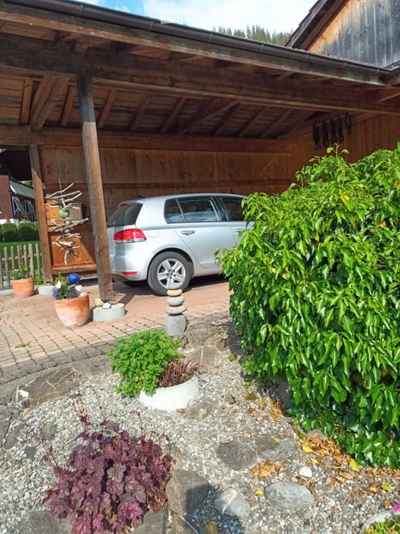 A covered wooden garage with a parked silver car underneath. The surrounding area features a small garden with potted plants, a stone stack, and a neatly trimmed hedge.