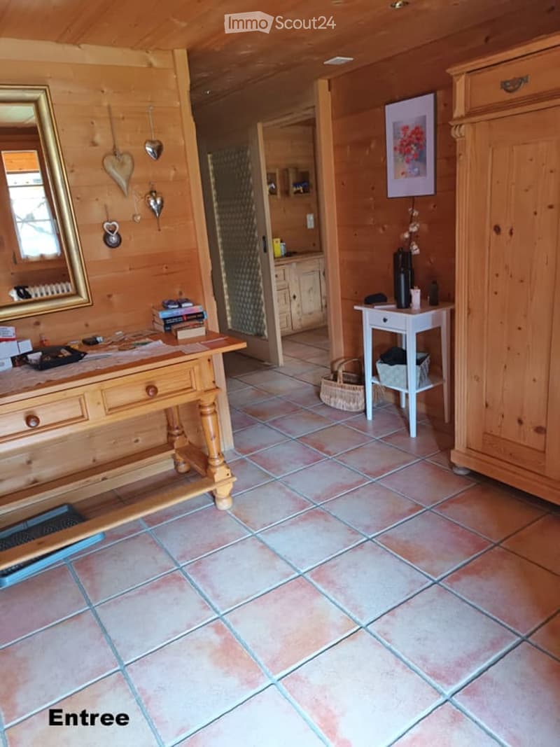 The image shows an entrance area with wooden walls and tiled flooring. Visible objects include a wooden table with books and a mirror, a small white table with a chair, a wooden cabinet, and a basket on the floor.
