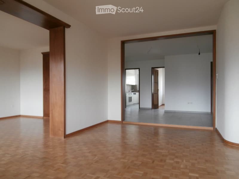 Empty room with wooden flooring and white walls, featuring an open doorway leading to another room with a kitchen area visible in the background.