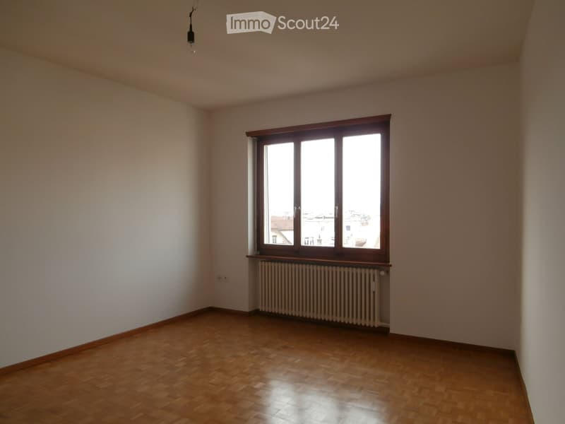 Empty room with wooden flooring, white walls, large window with wooden frame, radiator below the window, and a single light fixture hanging from the ceiling.