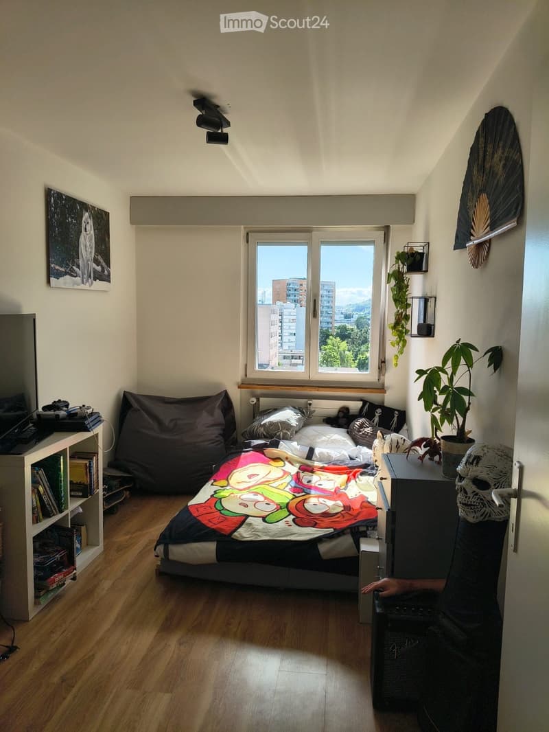 Cozy bedroom with wooden flooring, large window offering a city view, a bed with a colorful cartoon-themed bedspread, a bookshelf, a potted plant, and a skull on a nightstand.