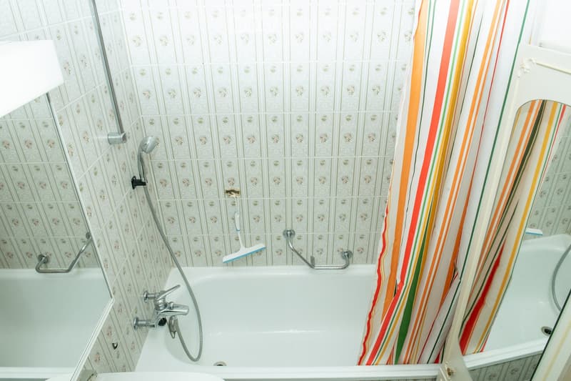 Bathroom with a white bathtub, multiple faucets, a colorful striped shower curtain, and a handheld showerhead with a handrail.
